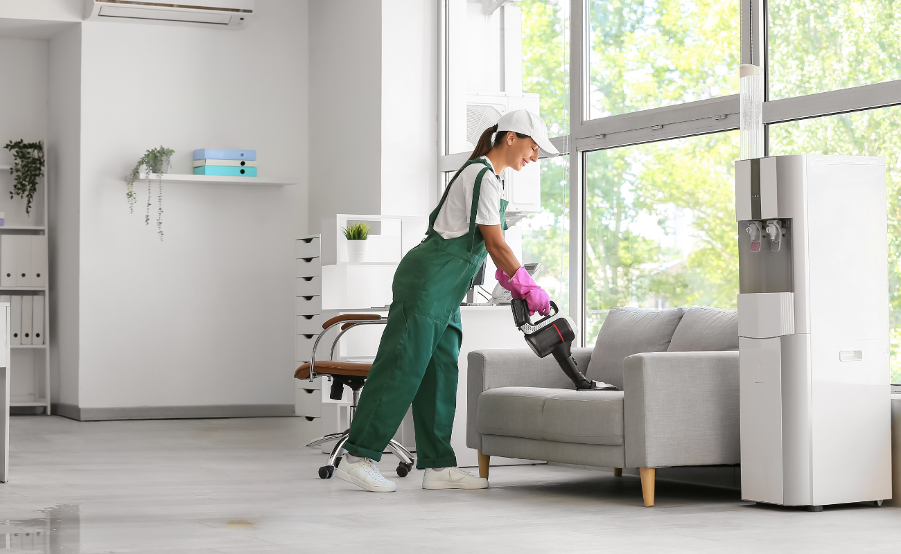 A woman wearing green overalls, a white cap, and pink gloves using a vacuum cleaner to clean a light gray sofa in an indoor space with large windows and a water dispenser.