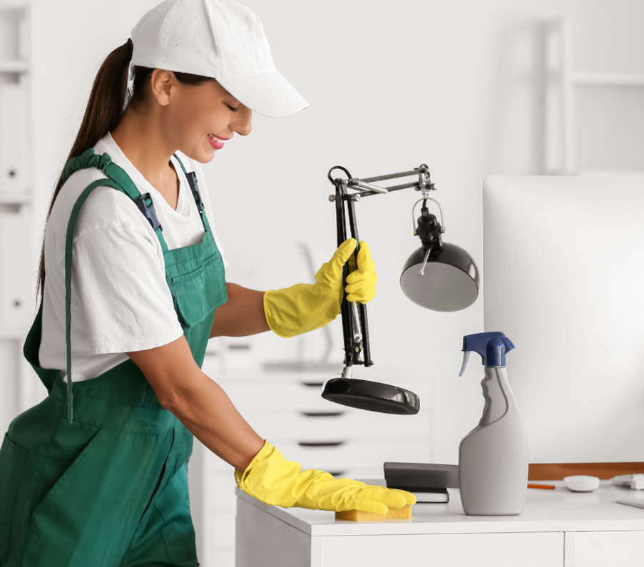 A woman cleaning a white desk with yellow rubber gloves, wearing a white cap, white t-shirt, and green apron, in a bright, organized office space.