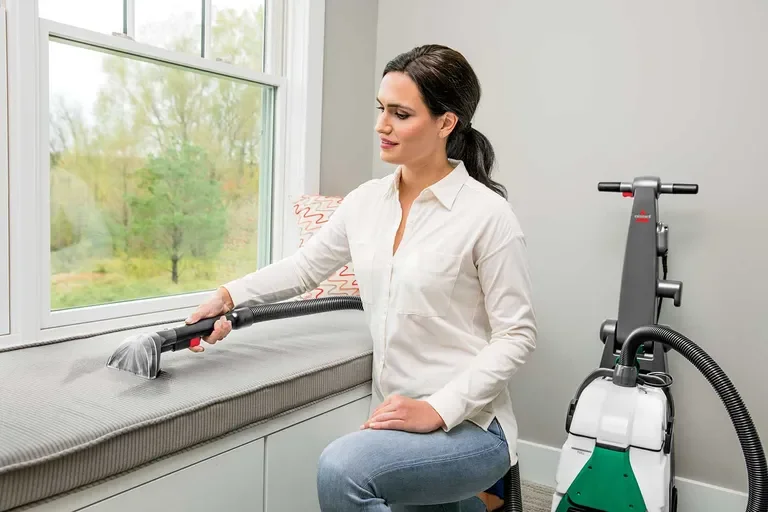 Woman cleaning a window with a handheld vacuum, sitting on a window seat in a bright room.