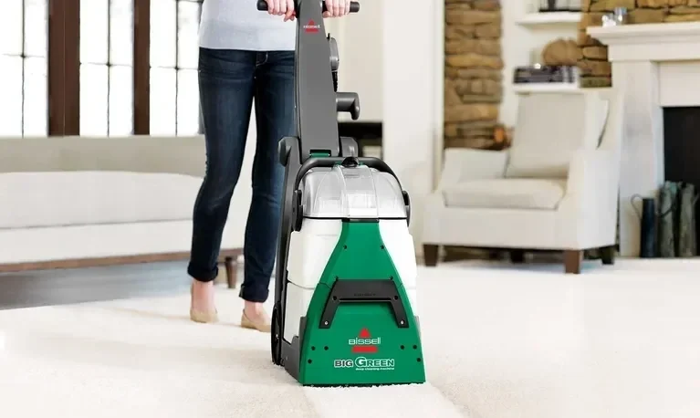 Person using a green and white Bissell Big Green carpet cleaner in a living room.
