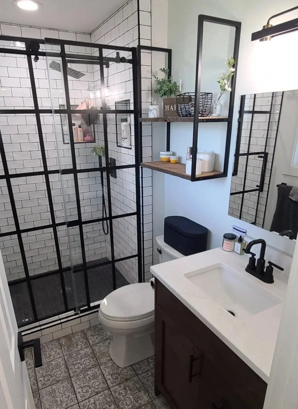 A modern bathroom with a walk-in shower enclosed by black-framed glass, white subway tile walls, a white toilet, a white vanity with a black faucet, and open shelving with decorative items and personal care products.